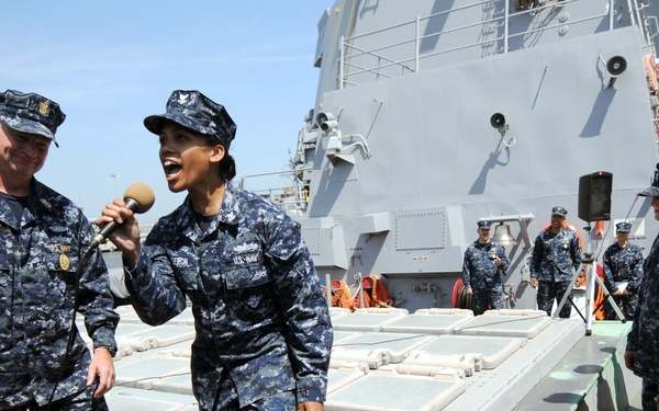 A group of U.S. Navy personnel in digital camouflage uniforms are gathered on the deck of a naval vessel. A female sailor is holding a microphone and appears to be speaking or performing, while a male sailor beside her reacts enthusiastically. Other sailors are lined up in the background observing the event.