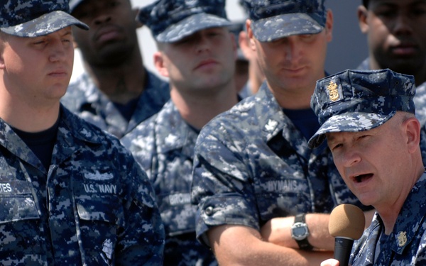 A senior U.S. Navy officer is addressing a group of enlisted sailors while holding a microphone. The personnel are all wearing the Navy Working Uniform (NWU) Type I, the blue digital camouflage pattern. The setting appears to be an outdoor military environment, likely on a ship's deck or base.