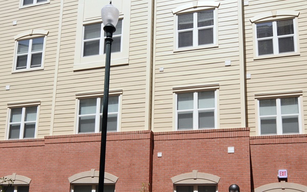 The image shows a multi-story residential or administrative building with red brick lower facade and beige siding upper floors. Four individuals, three wearing Navy Working Uniform (NWU) camouflage and one in civilian attire, are gathered near the building entrance. The scene appears to be an inspection or tour of a newly constructed or renovated military facility.