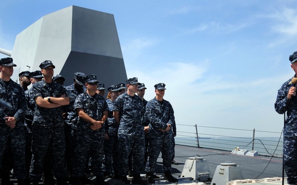A senior U.S. Navy officer addresses a group of sailors assembled on the deck of a naval warship. The officer, holding a microphone, appears to be giving a speech or briefing to the crew who are standing at ease in their Navy Working Uniform (NWU) digital camouflage. In the foreground, a Vertical Launch System (VLS) missile battery is prominently visible, indicating this is likely a destroyer or cruiser.