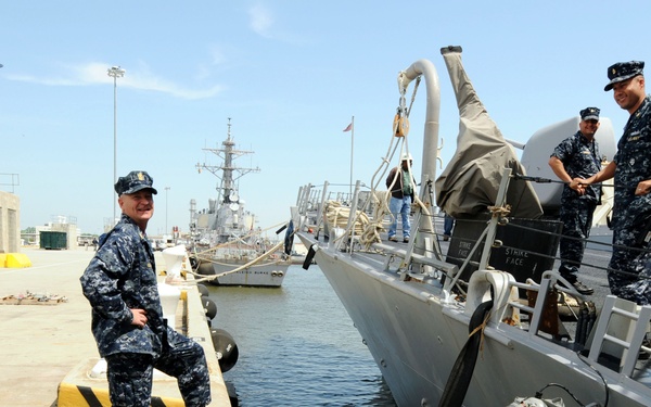 Several U.S. Navy personnel in blue digital camouflage (NWU Type I) uniforms are gathered at a naval pier alongside a gray military vessel. Two sailors stand on the dock conversing with crew members aboard the ship, suggesting an inspection, visit, or briefing. Additional naval vessels are visible in the background at what appears to be a U.S. Navy installation.