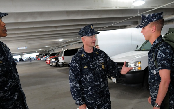 Three U.S. Navy personnel in Navy Working Uniform (NWU) Type I blue digital camouflage are conversing in what appears to be a parking garage or covered vehicle bay. The central figure, whose name tape reads 'WEST,' is holding a white hard hat and appears to be speaking to the sailor on the right, who is carrying a large olive drab duffel bag. The sailor on the left has a name tape reading 'WILLIAMS.'