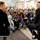 Two men seated on stools face each other in what appears to be a panel discussion or interview format in a modern indoor venue, likely a university or corporate cafeteria. A large audience of approximately 40-50 people, predominantly young adults, is seated in chairs watching the exchange. The setting suggests a formal but casual Q&amp;A or fireside chat event.