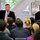 Two men stand at the front of a room addressing a seated audience in what appears to be a professional or corporate presentation setting. The man on the left, wearing a dark pinstripe suit with a red tie, is speaking into a microphone with an animated hand gesture. The man on the right, in a dark jacket, stands quietly beside him observing the audience.