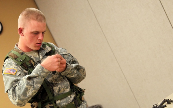 A U.S. Army soldier in ACU (Army Combat Uniform) is adjusting tactical gear or a vest in what appears to be an indoor training or classroom environment. A table in the foreground displays multiple disassembled firearms and weapon components. The setting suggests a weapons familiarization or equipment training exercise.