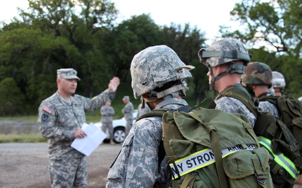 A military briefing or formation is taking place outdoors, with a soldier in ACU (Army Combat Uniform) without helmet addressing a group of fully equipped soldiers wearing helmets and carrying large rucksacks. The soldiers in formation have their backs to the camera and are wearing reflective safety bands on their packs, with text visible reading 'ARMY STRONG' and 'ARMY RESERVE'. The setting appears to be a paved area near a wooded location, likely a training exercise or pre-mission briefing.