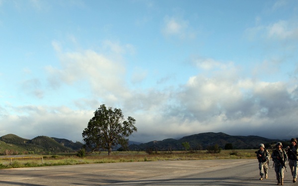 Three military personnel in combat uniforms and helmets are walking along a paved road or airstrip in what appears to be a training area. They are carrying weapons and moving in a group across an open, flat surface with rolling hills and trees visible in the background. The scene appears to be captured during early morning or late afternoon based on the low-angle lighting and long shadows.