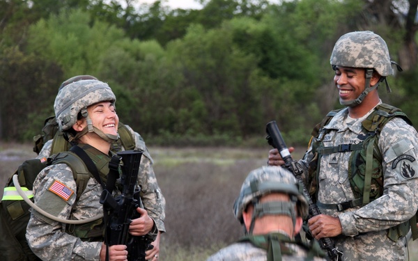 Several U.S. Army soldiers in ACU (Army Combat Uniform) digital camouflage are gathered in an outdoor field setting near a tree line. Two soldiers in the foreground are smiling and appear to be in a relaxed, candid moment during what appears to be a training exercise. The soldiers are fully equipped with helmets, body armor, rucksacks, and weapons.
