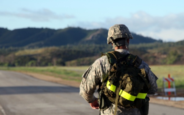 A soldier in US Army ACU (Army Combat Uniform) digital camouflage is walking or marching along a paved road while carrying a military rucksack with a high-visibility yellow reflective band. The setting appears to be a military installation or training area, with open fields, hills, and mountains visible in the background. At least one additional soldier is visible further down the road, suggesting a group ruck march or physical training event.