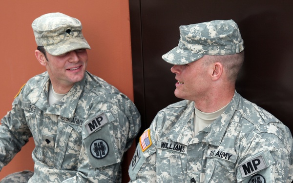 Two U.S. Army Military Police soldiers are seated and engaged in conversation. Both wear Army Combat Uniforms (ACU) with MP patches, unit insignia, and name tapes visible. The soldier on the right is identified as 'Williams' with the rank of Specialist, while the soldier on the left has a partially visible name tape.