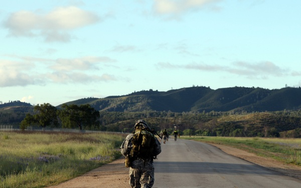 A group of military personnel in full combat gear are conducting a road march along a paved two-lane road in a rural, hilly landscape. The nearest soldier is prominently visible from behind, carrying a heavy rucksack and wearing a combat helmet and camouflage uniform. Additional soldiers are visible further down the road, suggesting a unit-level tactical movement or training exercise.
