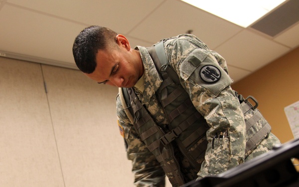 A U.S. Army soldier in ACU (Army Combat Uniform) with tactical vest is leaning over a table examining disassembled weapon components in what appears to be an indoor classroom or briefing room. The soldier is focused on the equipment laid out on the table in front of him. A map is visible on the wall in the background.