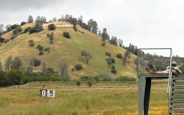 Two military personnel in camouflage uniforms are positioned on an elevated metal observation/firing platform at what appears to be an outdoor shooting range. The range features target boards visible in the middle distance across an open grassy field. A prominent rounded hill with scattered trees forms the backdrop under an overcast sky.