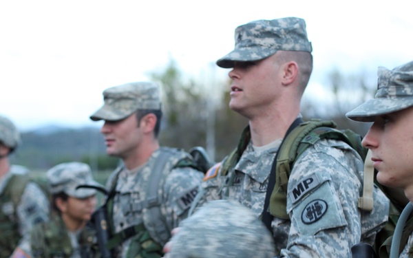 A group of U.S. Army soldiers standing in formation, equipped with full combat gear including rifles, backpacks, and ACU (Army Combat Uniform) camouflage. The central figure prominently displays 'MP' (Military Police) insignia on his uniform along with a unit patch. The setting appears to be an outdoor staging area or parking lot during overcast conditions.
