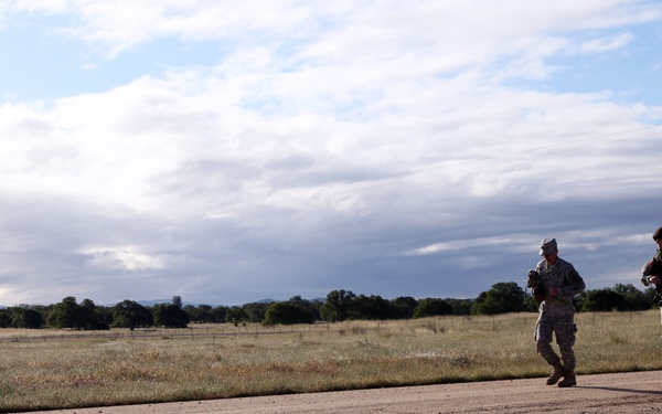 Two individuals in military camouflage uniforms are walking or moving along a dirt road or path in an open rural landscape. They appear to be carrying gear or equipment, with one person wearing a backpack. The setting is a wide, flat grassland area with trees visible on the horizon under a partly cloudy sky.