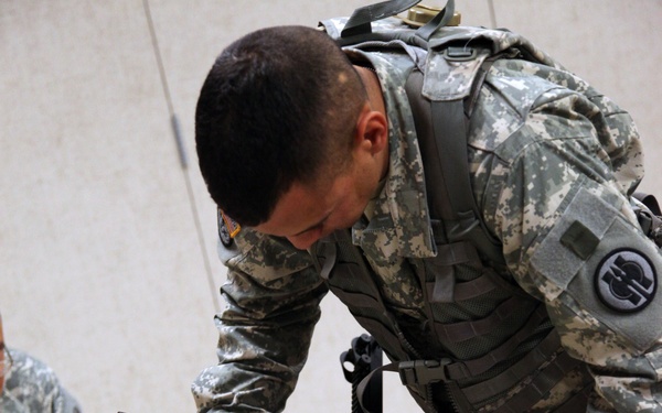 A U.S. Army soldier in ACU (Army Combat Uniform) with tactical vest and backpack is leaning over a white table performing a field strip or reassembly of a weapon, likely an M249 SAW or similar firearm. Multiple disassembled weapon components are spread across the table surface. A second soldier in ACU is partially visible on the left side of the frame.