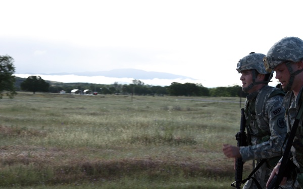 Two U.S. Army Military Police soldiers in ACU (Army Combat Uniform) are moving at a quick pace along a dirt road in what appears to be a training exercise or field operation. Both soldiers are wearing ACH helmets, carrying rifles, and bearing rucksacks. The setting is an open rural or training area with sparse vegetation and an overcast sky.