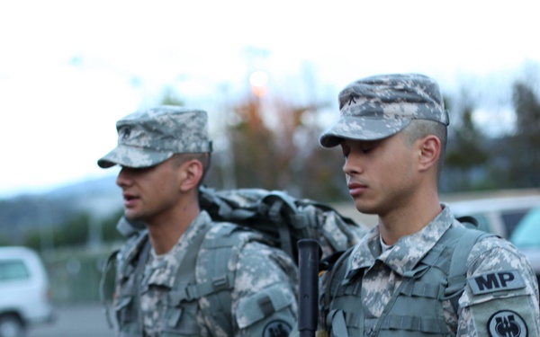 Two U.S. Army soldiers in Army Combat Uniform (ACU) digital camouflage pattern are standing at attention or in a formation outdoors. The soldier in the foreground displays an 'MP' (Military Police) tab and a unit patch on his left shoulder, and is holding an M16/M4-style rifle. Both soldiers are wearing plate carrier vests and patrol caps.