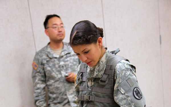 Two U.S. Army soldiers in ACU (Army Combat Uniform) digital camouflage are in an indoor training environment. A female soldier wearing a tactical vest is leaning over a table examining disassembled weapons, while a male soldier stands in the background observing. The setting appears to be a weapons familiarization or qualification training exercise.