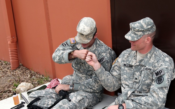 Two U.S. Army Military Police soldiers in ACU (Army Combat Uniform) are seated on a concrete surface against a building wall. The soldier on the right, identified by nametape as 'Williams,' holds the rank of Sergeant and wears an MP patch. Both soldiers appear to be taking a break and handling equipment, with weapons and gear visible on the ground beside them.