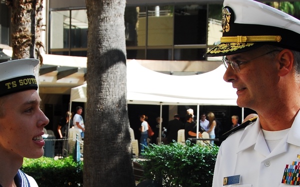 Two naval personnel are engaged in conversation in an outdoor urban setting. On the left is a young Australian Navy Cadet wearing a traditional sailor uniform with the cap inscription 'TS SOUT[H...]' and an 'Australian Navy Cadets' patch. On the right is a senior US Navy officer in white service dress uniform adorned with multiple ribbons, medals, and gold insignia.