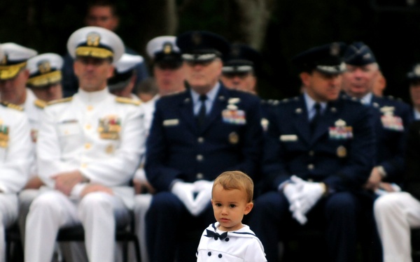 A toddler dressed in a miniature white sailor outfit walks across a brick plaza in front of a row of seated military officers in formal dress uniforms. The officers represent multiple branches of the U.S. military, including the Navy, Air Force, and Marine Corps. The contrast between the small child and the formally uniformed adults creates a visually striking and charming composition.