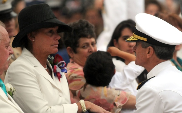 A military officer in white dress uniform is presenting a folded American flag to a woman dressed in white with a black wide-brimmed hat, consistent with a military funeral or memorial ceremony. A man in a white suit sits beside her, and a crowd of attendees is visible in the background. The solemn expressions and ceremonial flag presentation indicate this is a formal military honors service.