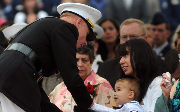 A military officer in dress uniform is presenting a folded American flag to a young woman holding a baby at what appears to be a military funeral or memorial ceremony. The baby is reaching out and touching the flag during the presentation. The scene is emotionally charged, with several attendees visibly grieving in the background.
