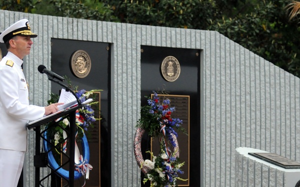 A naval officer in white dress uniform delivers a speech at a podium during what appears to be a military memorial ceremony. The setting features a memorial wall with bronze plaques and patriotic floral wreaths. A small toddler dressed in a white sailor outfit walks near a monument in the background.