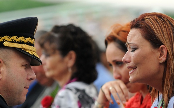 A military officer in dress uniform with white gloves appears to be presenting or offering condolences to a visibly emotional woman with red hair, who is wearing a corsage with red, white, and blue ribbons. Several other women are seated nearby, some also appearing emotional, suggesting this is a memorial or military ceremony. The scene is consistent with a formal military honors event, possibly a funeral or memorial service.