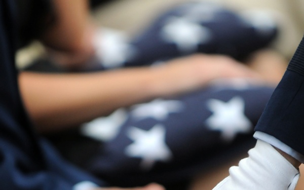 The image depicts a military funeral or memorial ceremony in which folded American flags are being presented to family members. A white-gloved hand, consistent with military dress uniform protocol, is placing or presenting a tri-folded U.S. flag onto the lap of a civilian recipient. Multiple recipients with folded flags are visible in the background, suggesting a group ceremony honoring multiple fallen service members.