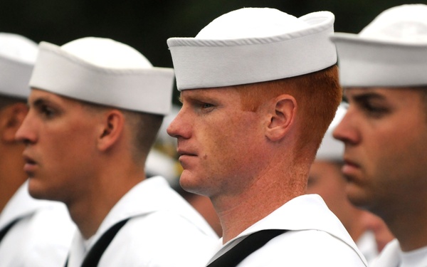 A group of U.S. Navy sailors in dress white uniforms are standing in formation, likely during a ceremony or parade. The sailors are wearing the distinctive white Dixie cup covers and black neckerchiefs. The central figure, a red-haired sailor, is in sharp focus and displays service ribbons and rating badges on his uniform.