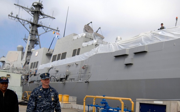 Two individuals walk along a naval pier beside a large gray warship docked at port. One person is wearing a U.S. Navy blue digital camouflage uniform, while the other is in civilian-style attire with a naval cap. The ship appears to be undergoing maintenance, evidenced by protective white coverings on portions of the vessel.