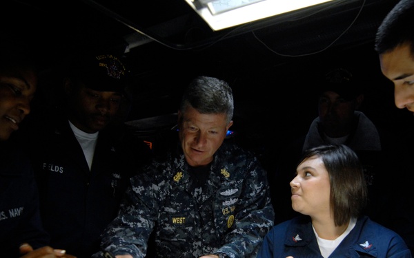 A group of U.S. Navy personnel are gathered around a table in a darkened space, appearing to review nautical charts or maps. A senior officer, whose name tape reads 'WEST,' is pointing at documents on the table while junior sailors observe. The setting appears to be a navigation or chart room aboard a naval vessel.