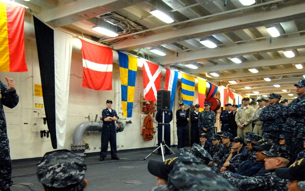 A senior U.S. Navy officer is addressing a large assembly of sailors in what appears to be a ship's hangar bay or interior deck space. The speaker is holding a microphone and speaking to a crowd of personnel seated on the floor and standing along the walls. Multiple nautical signal flags are displayed in the background, suggesting a formal all-hands or briefing event.