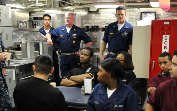 A U.S. Navy officer in digital camouflage uniform appears to be addressing a group of sailors in what looks like a ship's galley or mess hall. The gathering includes personnel in various Navy uniforms seated around tables and standing along the walls. The setting suggests an informal briefing or command interaction session aboard a naval vessel.