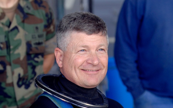A middle-aged man with gray-streaked hair is smiling and wearing a black XCEL wetsuit with blue harness straps and a dry suit neck ring. He is holding a yellow diving helmet with attached regulators and hardware. The setting appears to be a pre-dive preparation area with other individuals visible in the background, one wearing military camouflage.