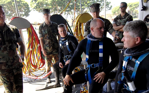 A group of military personnel in camouflage uniforms are gathered on what appears to be a dock or vessel platform. Three individuals are dressed in black wetsuits with blue diving harnesses and equipment, appearing to be preparing for or debriefing after a dive operation. Several uniformed soldiers stand nearby observing or providing instruction.