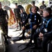 A group of military personnel in camouflage uniforms are gathered on what appears to be a dock or vessel platform. Three individuals are dressed in black wetsuits with blue diving harnesses and equipment, appearing to be preparing for or debriefing after a dive operation. Several uniformed soldiers stand nearby observing or providing instruction.