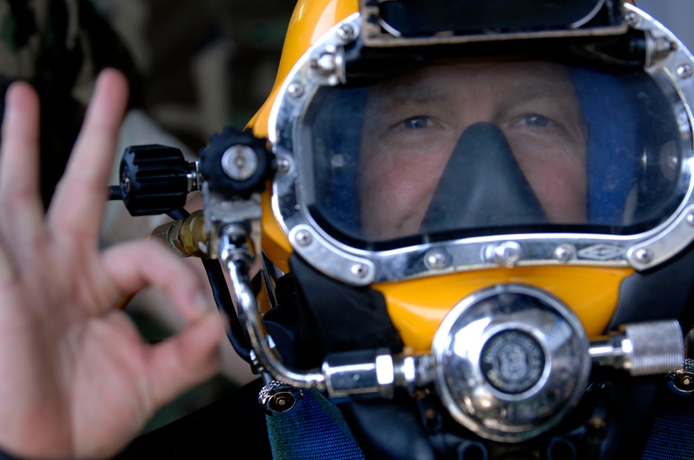 A close-up photograph of a diver wearing a professional full-face diving helmet, predominantly yellow with a large clear visor and chrome/metal fittings. The diver is making an 'OK' hand signal with their left hand in the foreground. The image is sharp and well-composed, focusing on the diver's face through the helmet visor.