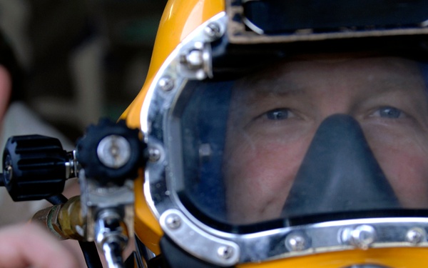 A close-up photograph of a diver wearing a professional full-face diving helmet, predominantly yellow with a large clear visor and chrome/metal fittings. The diver is making an 'OK' hand signal with their left hand in the foreground. The image is sharp and well-composed, focusing on the diver's face through the helmet visor.