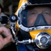 A close-up photograph of a diver wearing a professional full-face diving helmet, predominantly yellow with a large clear visor and chrome/metal fittings. The diver is making an 'OK' hand signal with their left hand in the foreground. The image is sharp and well-composed, focusing on the diver's face through the helmet visor.