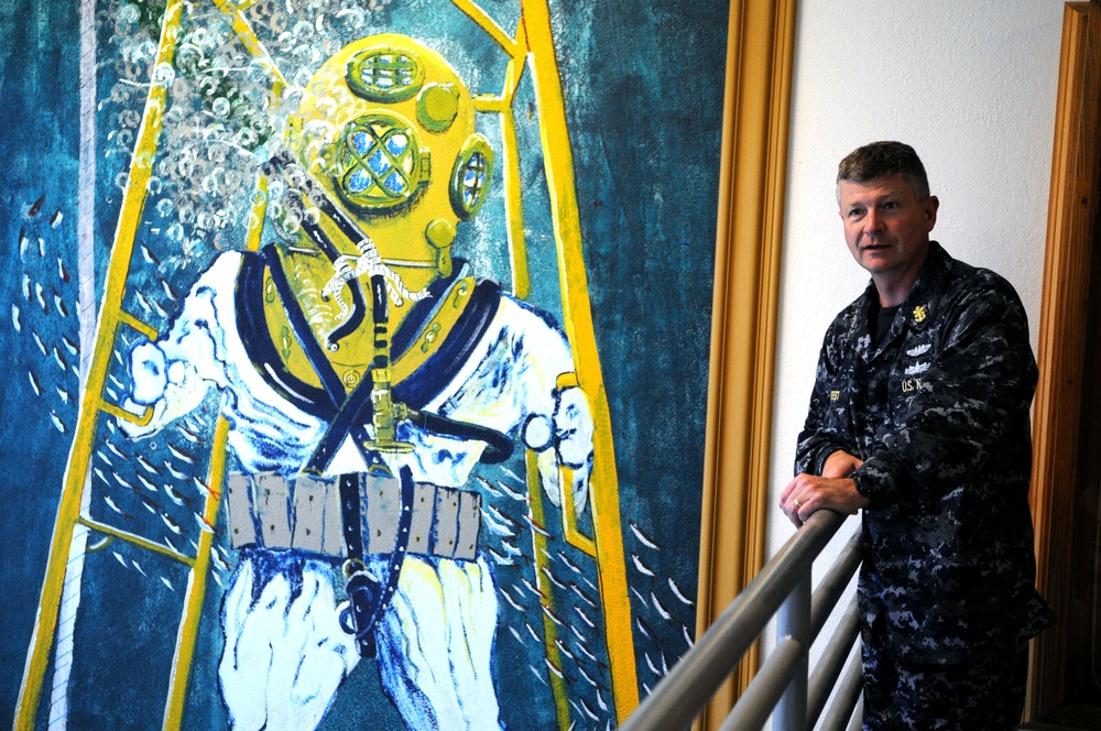 A U.S. Navy servicemember in digital camouflage (NWU Type I) uniform stands beside a large painted mural depicting a classic deep-sea diver in a traditional Mark V diving helmet and suit. The mural is rendered in bold blues, yellows, and whites, showing the diver ascending a yellow ladder underwater. The setting appears to be an interior stairwell or hallway of a naval facility.