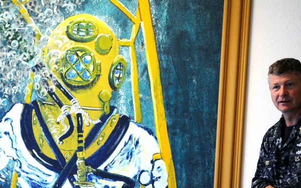 A U.S. Navy servicemember in digital camouflage (NWU Type I) uniform stands beside a large painted mural depicting a classic deep-sea diver in a traditional Mark V diving helmet and suit. The mural is rendered in bold blues, yellows, and whites, showing the diver ascending a yellow ladder underwater. The setting appears to be an interior stairwell or hallway of a naval facility.