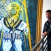 A U.S. Navy servicemember in digital camouflage (NWU Type I) uniform stands beside a large painted mural depicting a classic deep-sea diver in a traditional Mark V diving helmet and suit. The mural is rendered in bold blues, yellows, and whites, showing the diver ascending a yellow ladder underwater. The setting appears to be an interior stairwell or hallway of a naval facility.
