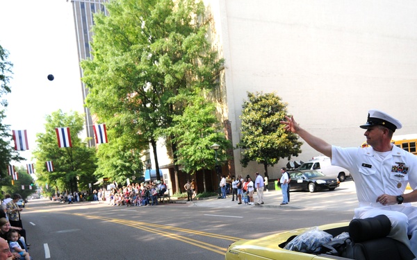 A military parade is taking place on a city street, featuring a naval officer in white dress uniform riding in a yellow convertible car marked as the 'Reviewing Officer.' The sign on the vehicle identifies the occupant as MCPON Rick West, Master Chief of the Navy. Crowds of spectators line the sidewalk on the left side of the street to watch the parade.
