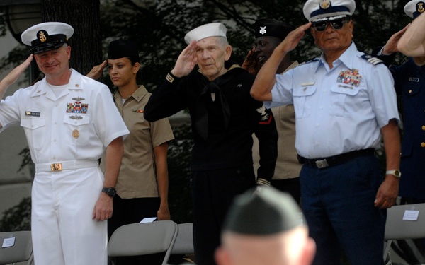 A formal military ceremony featuring personnel from multiple branches of the U.S. Armed Forces standing on a raised platform and rendering salutes. The scene includes officers and enlisted members in various dress uniforms representing the Army, Navy, Air Force, and Marine Corps. An audience of uniformed personnel in white uniforms is visible in the foreground, facing the platform.
