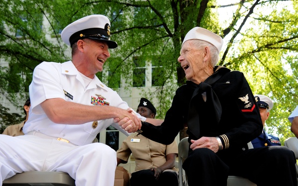 A U.S. Navy officer in white dress uniform shakes hands and shares a laugh with an elderly veteran wearing a dark Navy enlisted uniform with a white Dixie cup hat. The scene appears to be an outdoor military ceremony or commemorative event, with multiple service members seated in the background. The interaction conveys warmth and mutual respect between generations of naval service members.