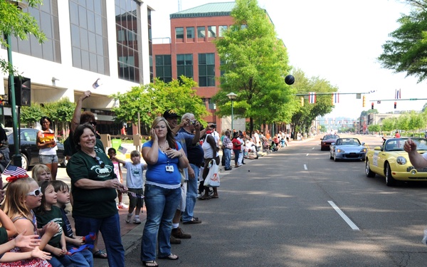 A U.S. Navy officer in dress whites is tossing items to spectators lining a street during a parade. Children and adults crowd the sidewalk reaching out to catch what appear to be small packaged items from a clear plastic bag. The parade takes place on a downtown urban street with buildings, trees, and vehicles visible in the background.
