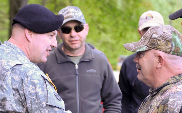 A U.S. Army soldier in ACU (Army Combat Uniform) with a black beret is shaking hands with a civilian man wearing Mossy Oak camouflage clothing and a camo cap. The interaction appears to be a formal or ceremonial greeting, taking place outdoors with several other individuals observing in the background. The setting appears to be a wooded or park-like area during spring or early summer based on the green foliage visible.